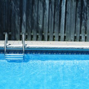 Clear blue swimming pool water with a metal ladder and wooden fence.